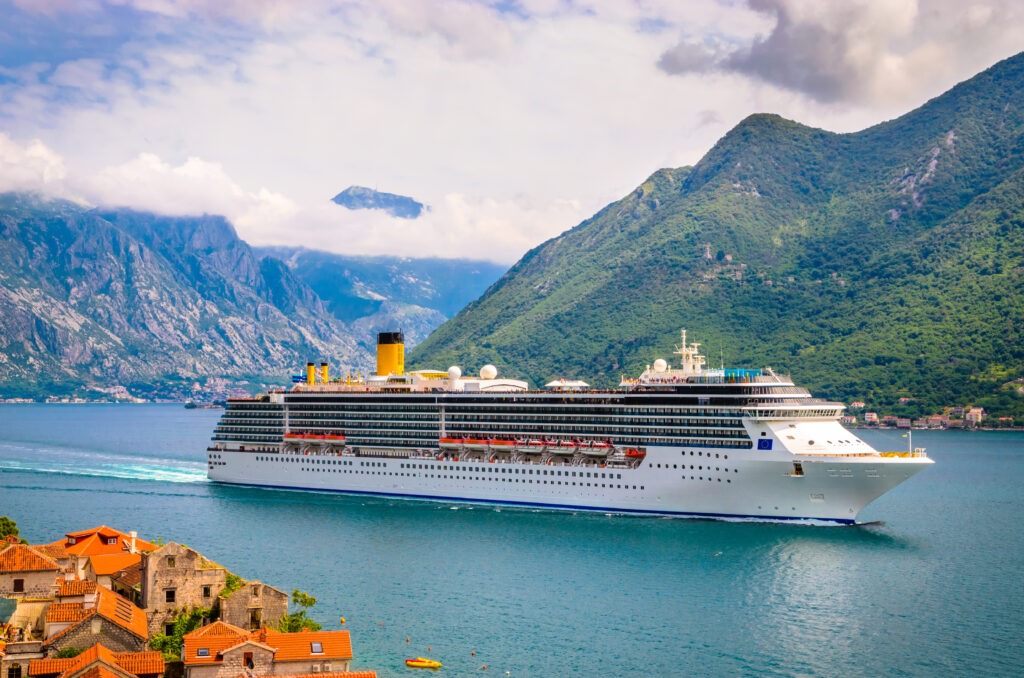 A large cruise ship docked near a coastal town, with towering mountains and cloudy skies in the background.