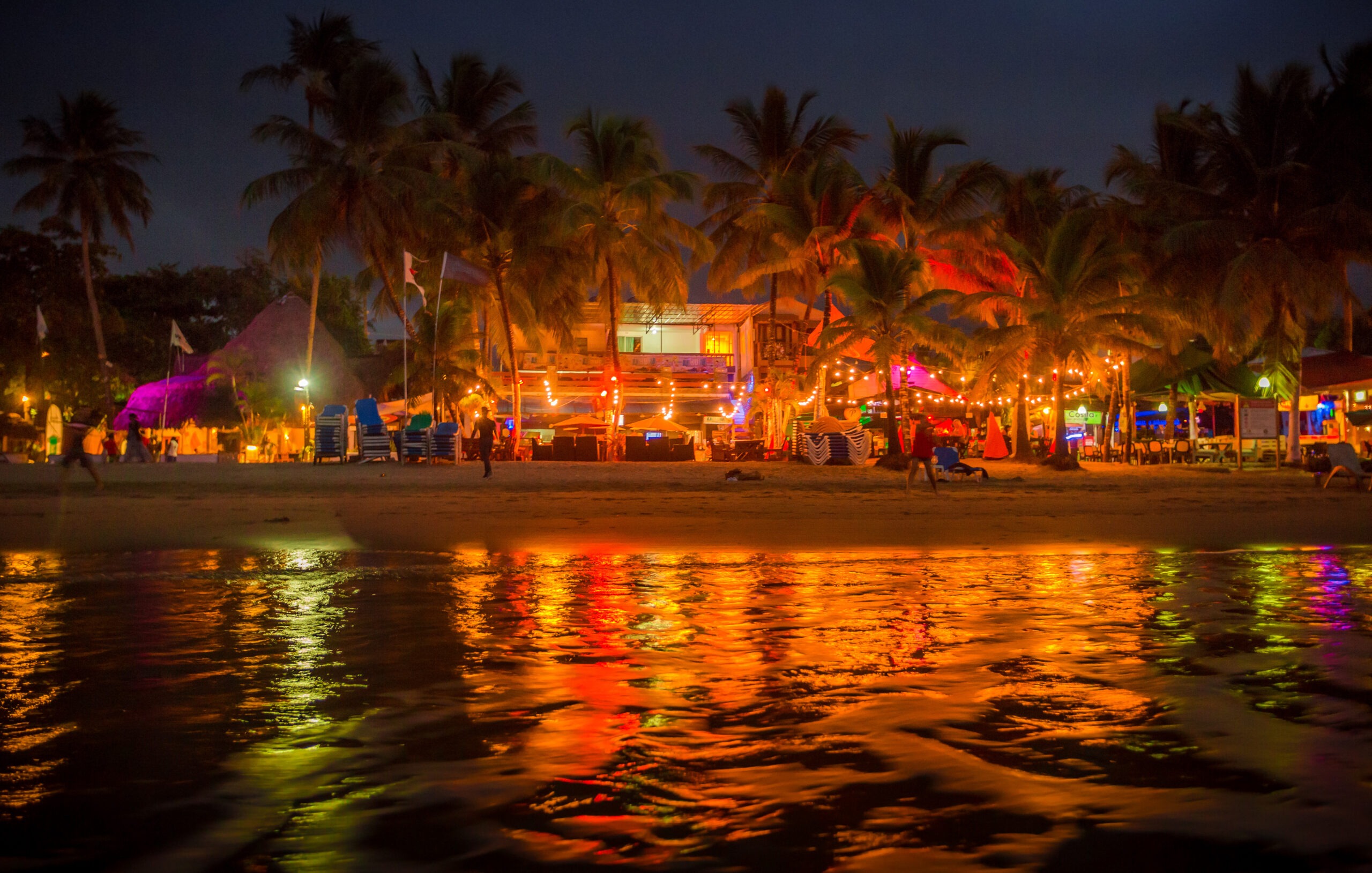 Nighttime tropical beach scene in Punta Cana with illuminated palm trees, a lively beachside venue, vibrant reflections on the water, and people strolling by.