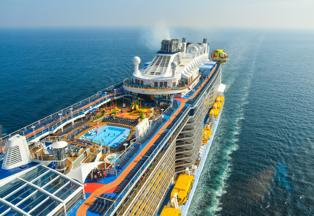 Aerial view of a vibrant cruise ship with colorful decks, a pool, parasailing activity, and the ocean stretching to the horizon.