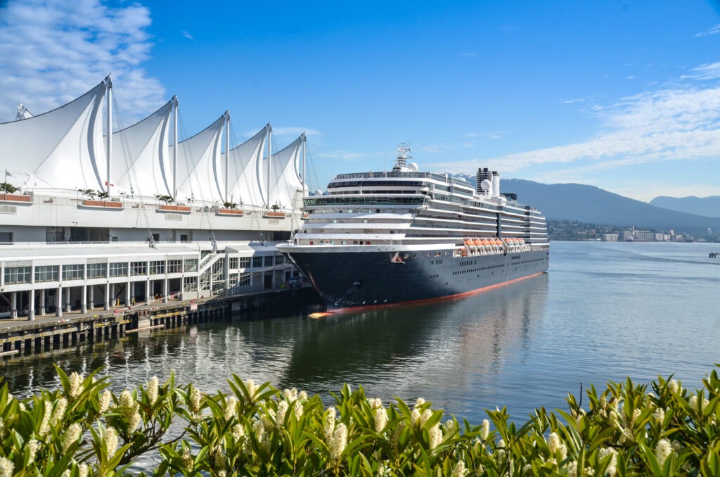 Cruise ship docked beside a modern building with sail-like roofs, calm waters in front, and distant mountains under a blue sky.