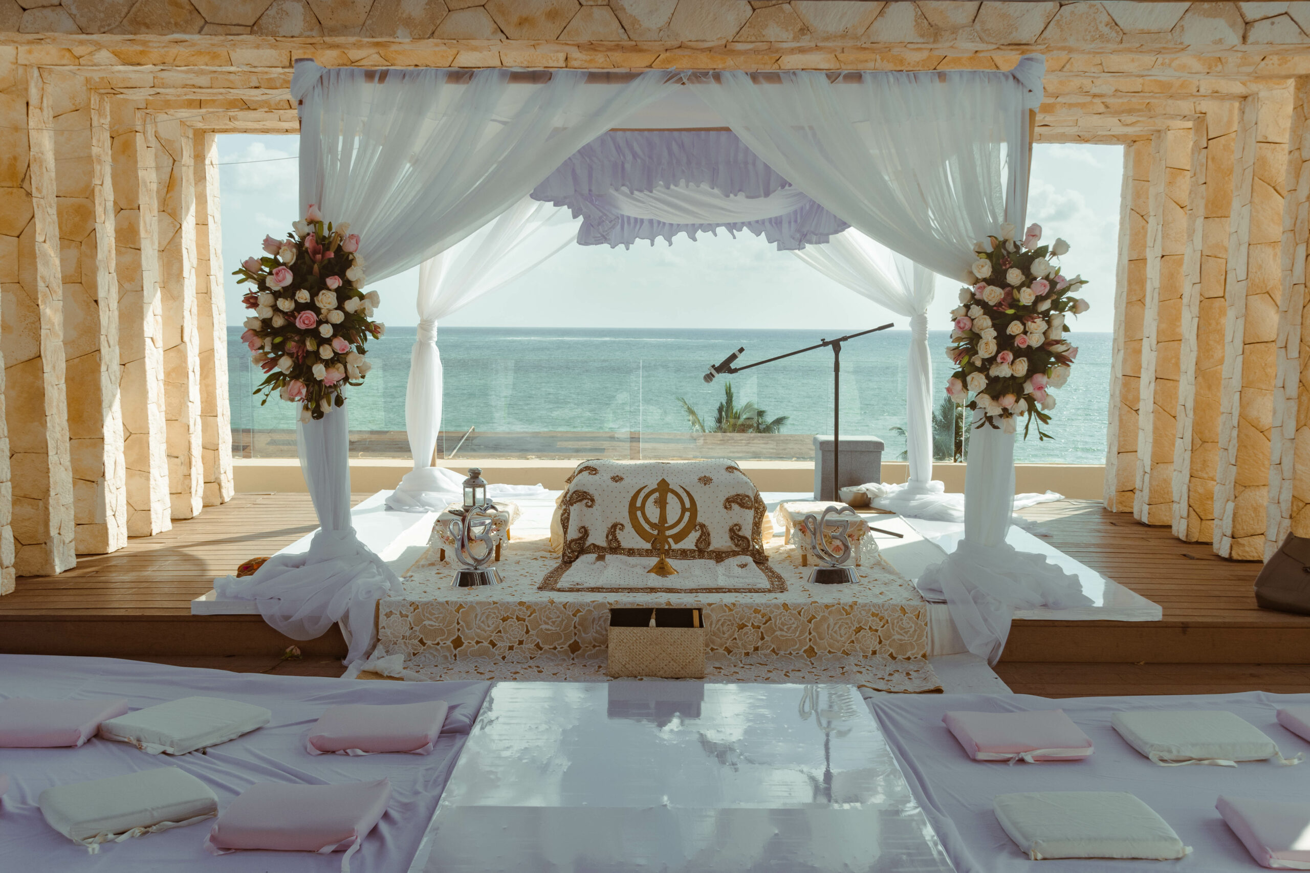 A Sikh wedding (Anand Karaj) setup with an ocean backdrop, featuring an ornate ceremonial seat, elegant drapery, and floral arrangements.