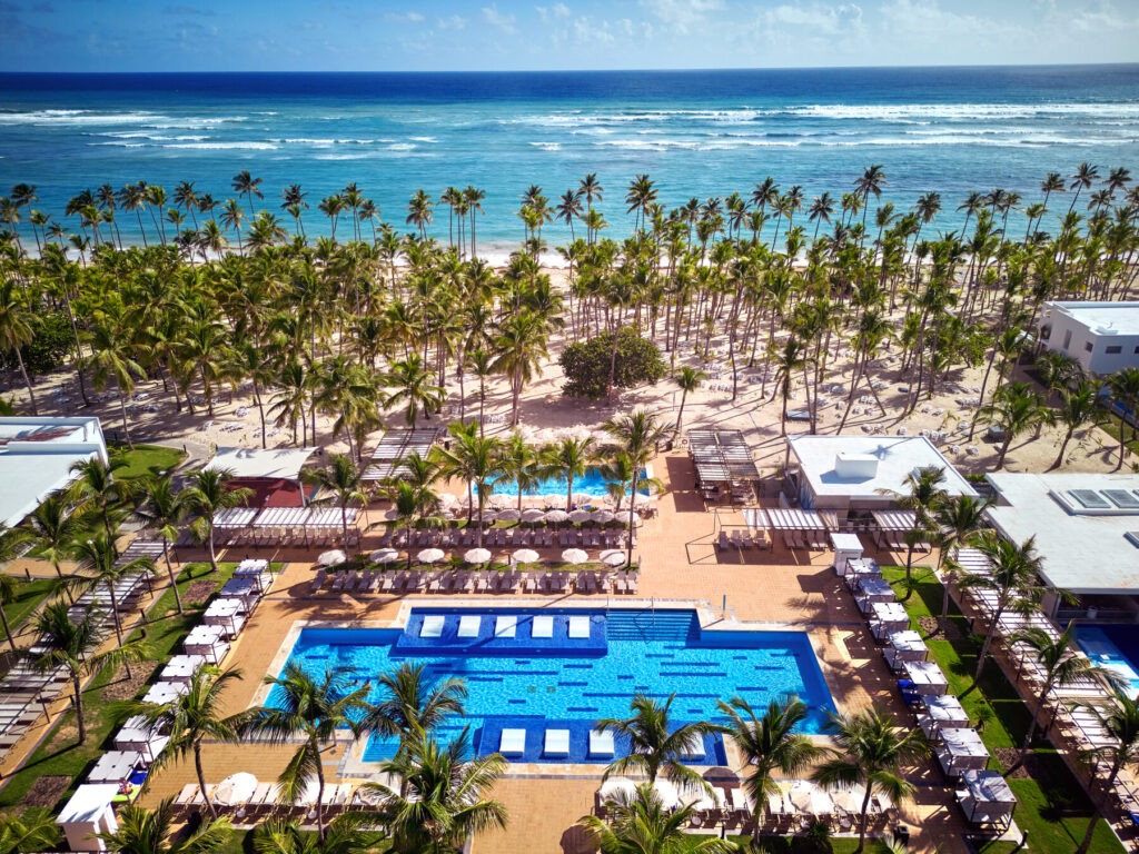 Aerial view of a luxurious beachfront resort in Punta Cana featuring a clear blue swimming pool, sun loungers, palm tree-lined sandy shores, and a vast turquoise ocean in the background.