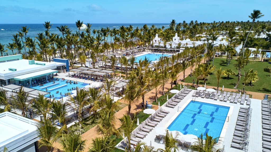 Aerial view of a tropical RIU Palace resort with palm trees, swimming pools, and beachfront facing the ocean.