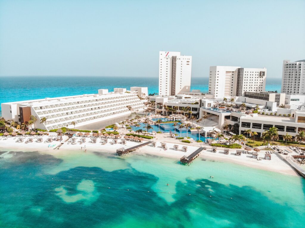 Aerial view of a Cancun hotel with white structures, turquoise pools, and a sandy beach by clear blue waters, ideal for bachelor and bachelorette parties.