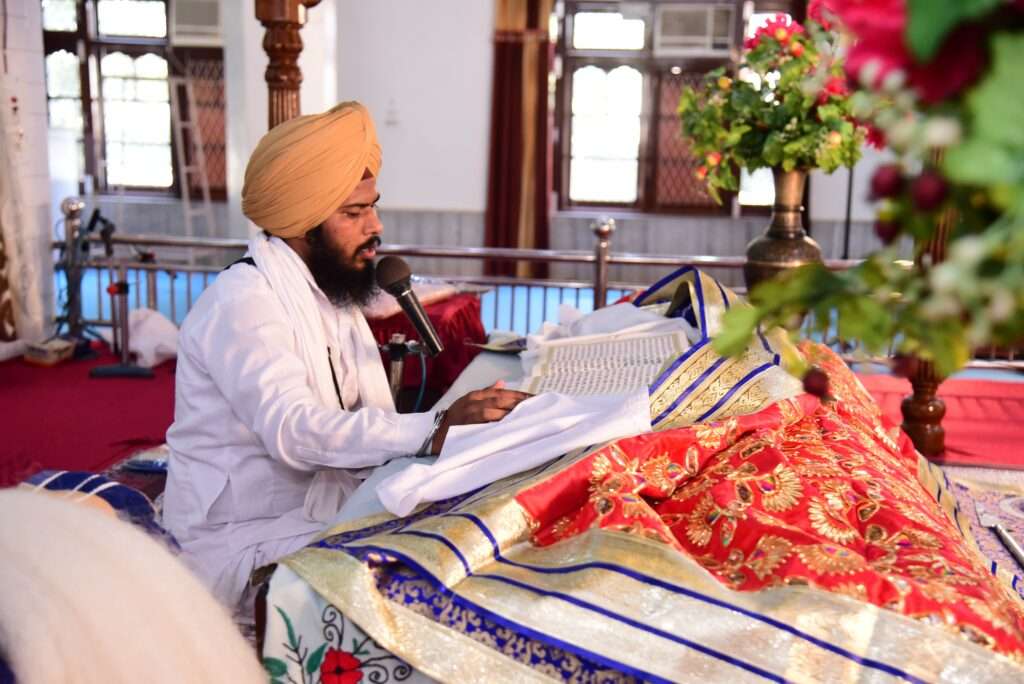 A Sikh man in traditional attire conducting an Anand Karaj (Sikh Wedding) beside the Guru Granth Sahib adorned with a vibrant cover admist the discussion of the wedding lehenga ban. 