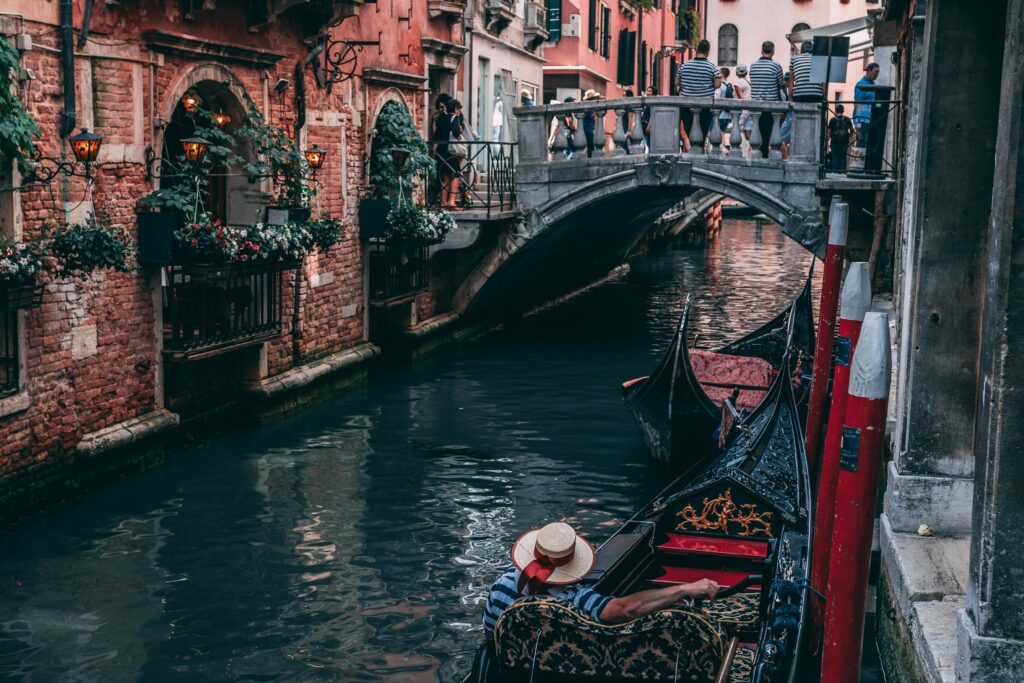 A gondolier in traditional attire navigates a gondola through a narrow canal in Venice, Italy, with a picturesque bridge and historic buildings adorned with flowers in the background.